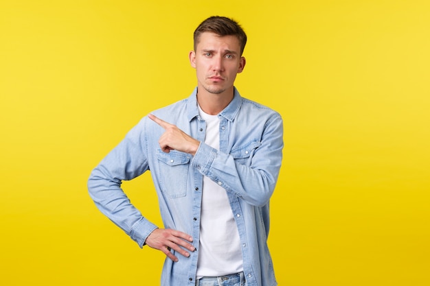 Skeptical arrogant blond handsome guy frowning and looking unbothered while pointing upper left corner at unamusing average promo banner, standing yellow background.