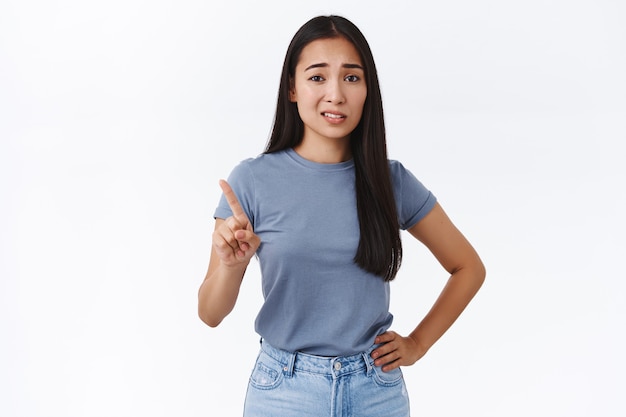 Skeptical and arrogant sassy young asian woman shaking finger in rejection, give refusal and looking with disdain or scorn, underestimate someone, standing white wall not giving any chance