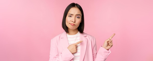 Skeptical asian businesswoman saleswoman sulking and looking with disapproval while pointing fingers right showing bad info upsetting news standing over pink background
