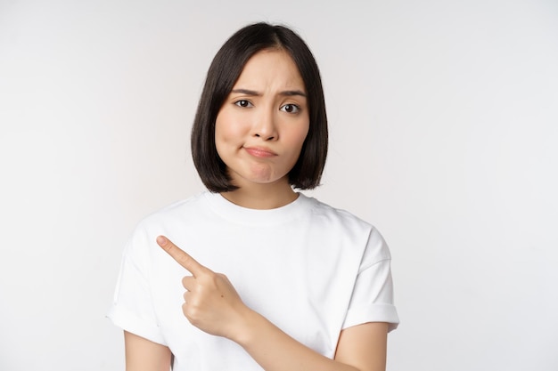 Скептически настроенная азиатка в белой футболке, указывающая на продукт или логотип с разочарованной гримасой, не любит и жалуется на что-то, стоящее на белом фоне