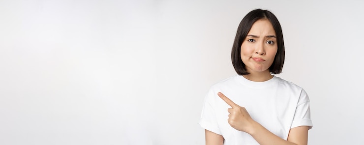 無料Photo 失望したしかめっ面で製品またはロゴを指している白いtシャツの懐疑的なアジアの女の子は嫌いで、白い背景の上に立っているsmthに文句を言う