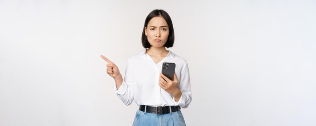 懐疑的なアジアの現代の女の子は、スマートフォンの人差し指を空のスペースに残し、躊躇している