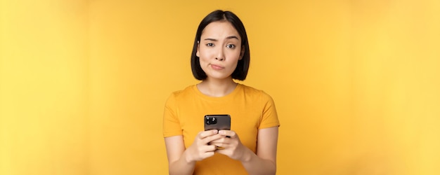 黄色の背景の上に立っているカメラを疑ってスマートフォンを保持している懐疑的なアジアの女性