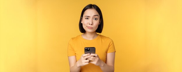 Skeptical asian woman holding smartphone looking with doubt at camera standing over yellow background
