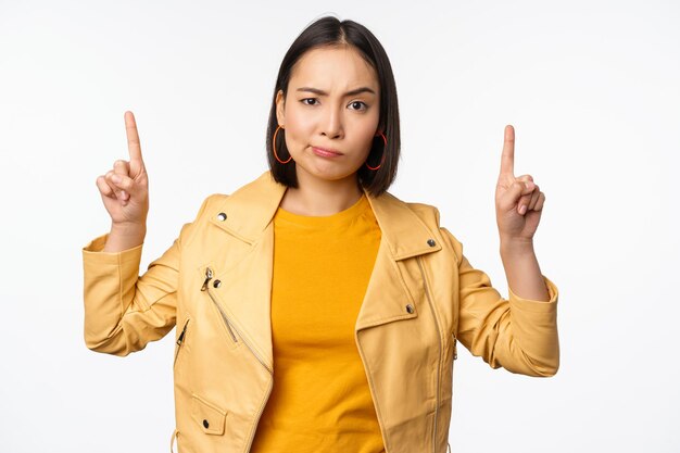 Skeptical asian woman pointing fingers up frowning and looking with disapproval complaining at smth standing doubtful against white background