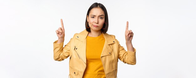 Skeptical asian woman pointing fingers up frowning and looking with disapproval complaining at smth standing doubtful against white background