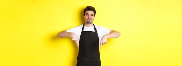 Skeptical barista in black apron pointing fingers down at bad product looking displeased and unamuse