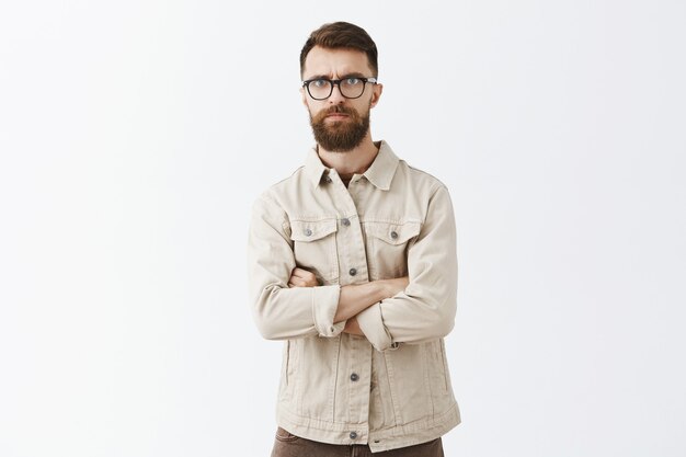 Skeptical bearded man in glasses posing against the white wall