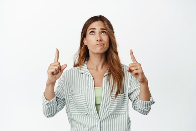 Skeptical brunette woman has concern, pointing fingers and looking up with sad smirking face and frown, disappointed in smth, unamused by advertisement, white background.