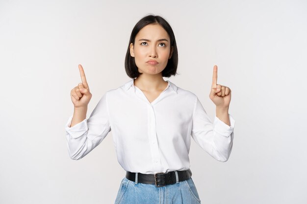 Skeptical businesswoman asian office manager pointing fingers up and grimacing doubtful hesitating standing over white background