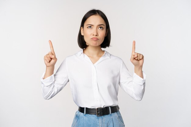 Скептически настроенная деловая женщина азиатский офис-менеджер показывает пальцем вверх и гримасничает, сомневаясь, стоя на белом фоне