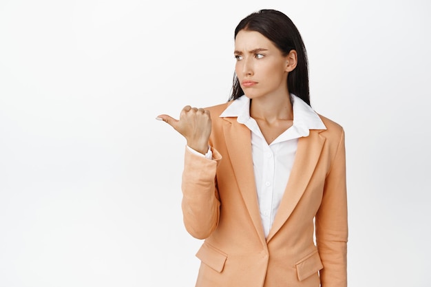 Free photo skeptical businesswoman pointing looking left with frowning face standing in suit against white background