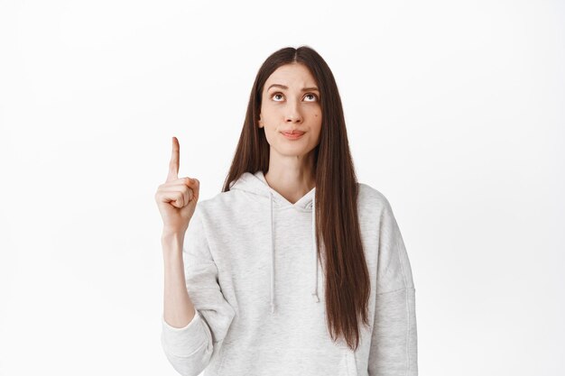Скептическая кавказская девушка с длинными волосами, хмурящаяся и сомневающаяся в продукте, указывая на пустое пространство сверху, показывая рекламу, стоя на белом фоне