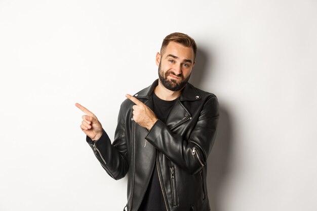 Skeptical and disapleased man in leather jacket, pointing fingers at upper left corner, showing bad promo offer, standing reluctant.