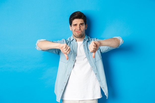 Free photo skeptical and disappointed 25 years old man in casual outfit dislike something bad, showing thumbs-down and grimacing displeased, standing against blue background.