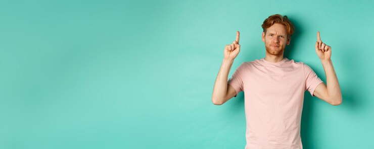 Free Photo skeptical and disappointed redhead man pointing fingers up frowning displeased and looking upset sta