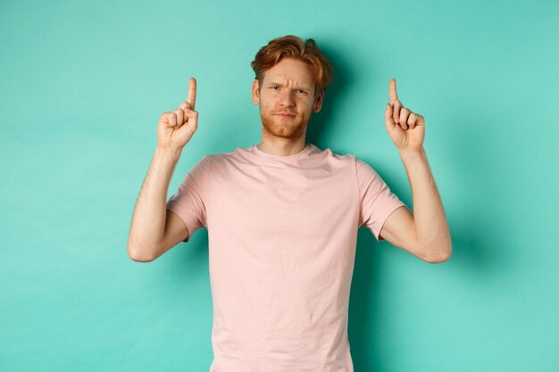 Skeptical and disappointed redhead man pointing fingers up, frowning displeased and looking upset, standing over turquoise background