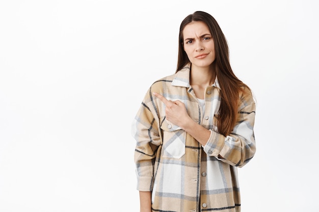 Skeptical or disappointed young woman grimacing with doubt face, pointing aside at something strange or doubtful, standing displeased against white wall