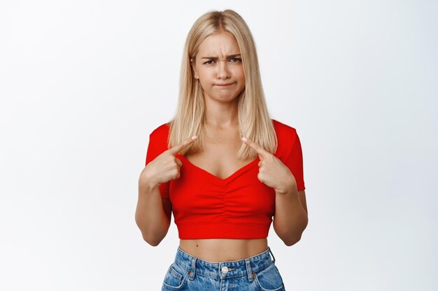 Skeptical and doubtful blond girl pointing fingers at herself grimacing and frowning upset standing unconfident against white background