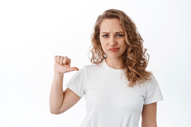 Skeptical and doubtful blond woman grimacing and frowning pointing at herself being unconfident standing over white background
