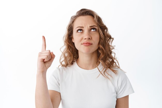 Skeptical and doubtful blond woman grimacing pointing and looking up unsure standing against white background Copy space
