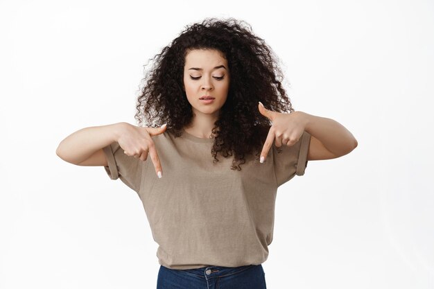 Skeptical and doubtful curly girl pointing fingers down, looking below with unsure face expression, standing in t-shirt against white background