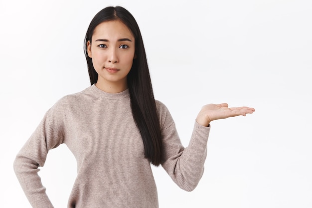 Skeptical good-looking asian woman in casual sweater, smirk unsure and unimpressed, present product, pointing right, holding something on blank white wall, standing studio advertising