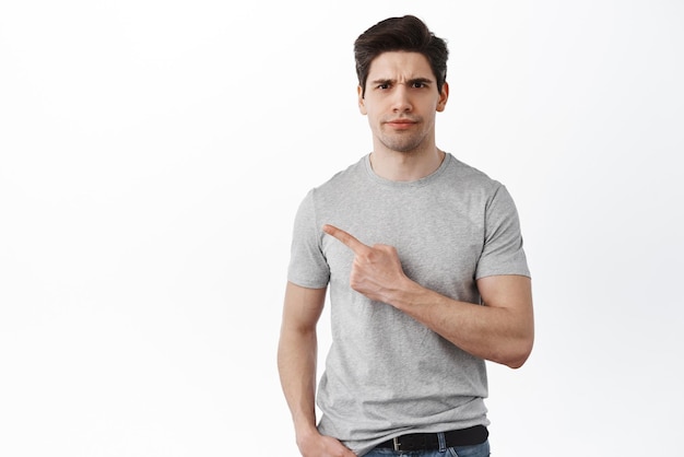 Skeptical guy frowns and points aside has doubts Disappointed young man complaining showing something strange standing confused over white background