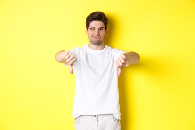 Skeptical guy showing thumbs down and smirking, dislike and disagree, standing over yellow background. Copy space