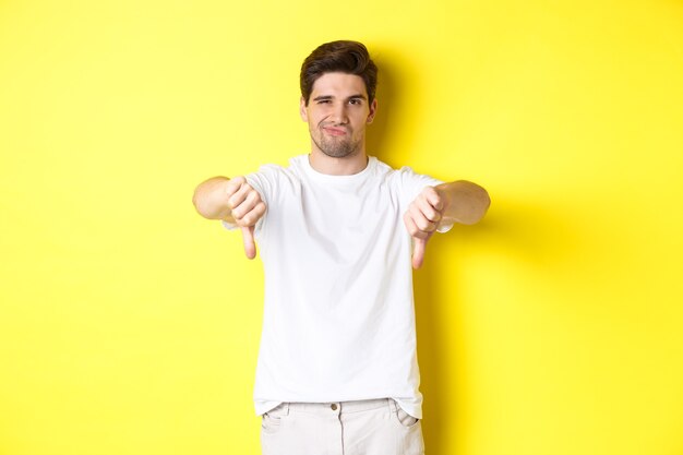 Skeptical guy showing thumbs down and smirking, dislike and disagree, standing over yellow background