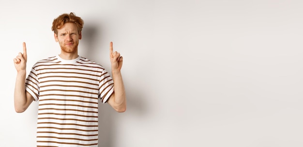 Skeptical handsome redhead man pointing fingers up grimacing and looking doubtful at camera standing