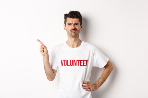 Skeptical and hesitant male volunteer in t-shirt grimacing doubtful, pointing finger left at promo offer, white background