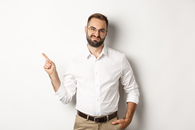 Skeptical male entrepreneur grimacing, pointing finger left with displeased face, complaining on