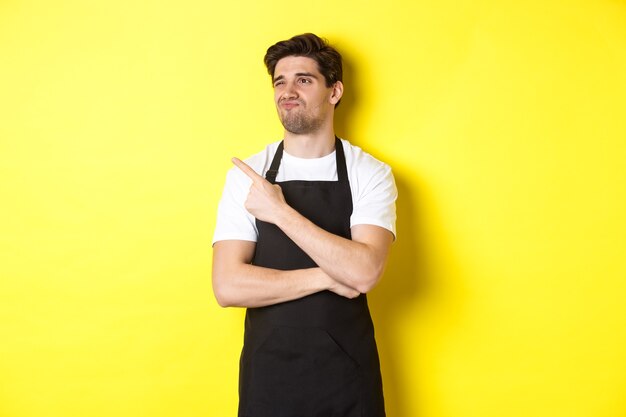 Skeptical male seller in black apron looking displeased, grimacing and pointing left at advertisement, standing over yellow background.