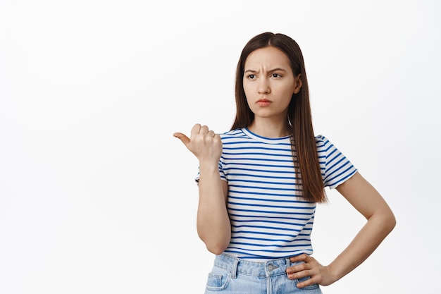 Ragazza scettica e schizzinosa che guarda, punta a sinistra con sopracciglia solcate e faccia giudicante, delusa, esprime incredulità o sentimenti sospetti, in piedi su sfondo bianco.