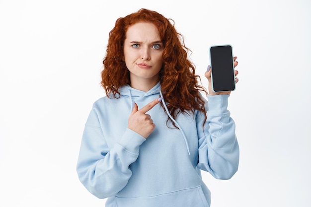 Skeptical redhead girl look confused, pointing at empty mobile phone screen with doubtful face, standing upset with app  on white