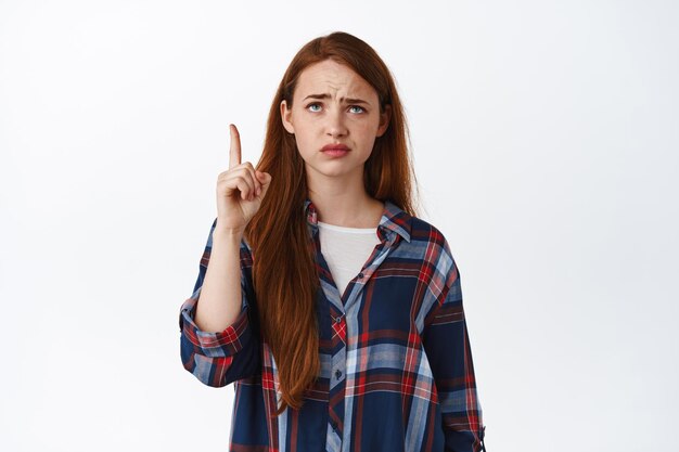 Skeptical redhead girl points at something lame, grimacing and frowning upset, pointing and looking with regret, unfair thing, standing over white background