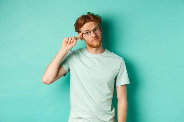 Skeptical redhead man scolding someone stupid or crazy, pointing finger at head and staring at camera, standing over mint background