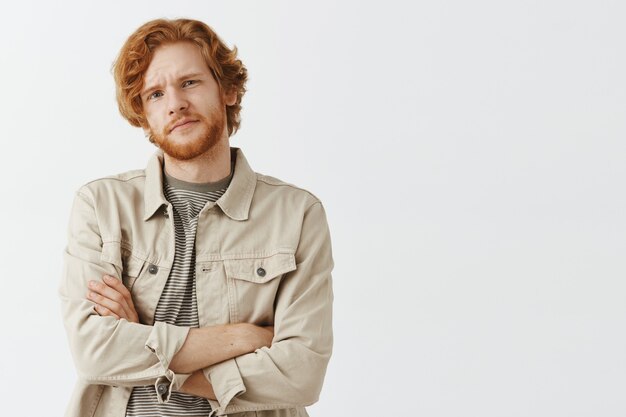 Skeptical and reluctant bearded redhead guy posing against the white wall