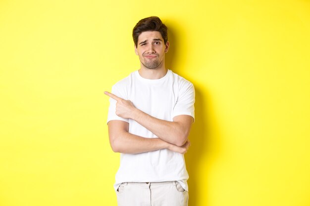 Skeptical and reluctant guy in white t-shirt, pointing finger left and smirking unamused, showing bad advertisement, standing over yellow background