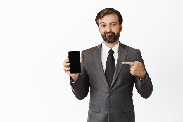 Skeptical salesman pointing finger at phone screen and grimacing disappointed negative opinion about app standing over white background