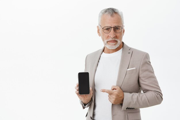 Skeptical and unamused old businessman pointing finger at smartphone with reluctant expression