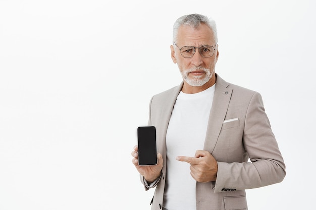 懐疑的で面白くない老人実業家が嫌な表情でスマートフォンに人差し指