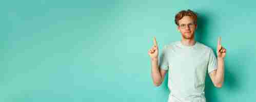 Free photo skeptical and unamused redhead man in glasses and tshirt pointing fingers up and showing something u