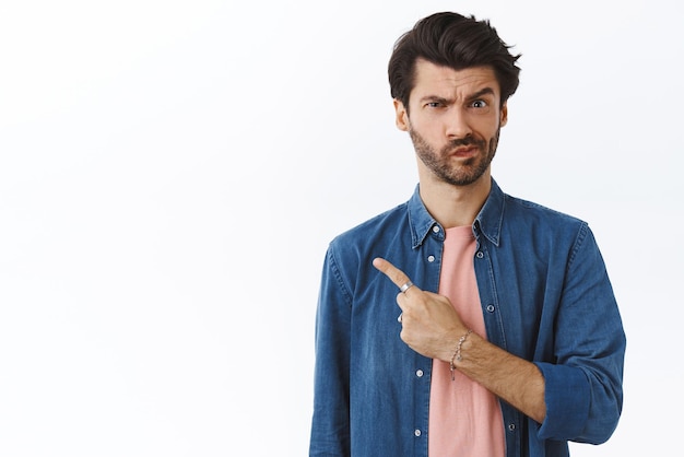 Foto gratuita scettico incerto e sospettoso bell'uomo di 25 anni con barba accigliata ghigno insicuro puntando a sinistra e fissando dubbioso la fotocamera non può decidere di rimanere indeciso su sfondo bianco