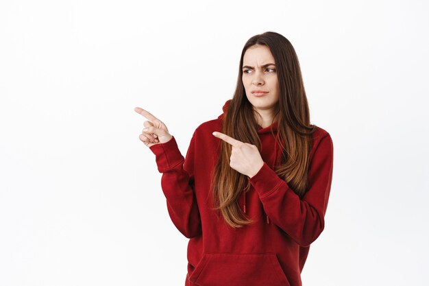 Skeptical woman tilt back and look with disdain or doubt at product promo deal, pointing and looking aside at copy space with something strange, standing against white wall