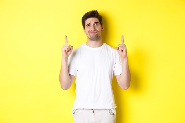 Skeptical young man pointing and looking up at something bad, judging offer, standing over yellow background.