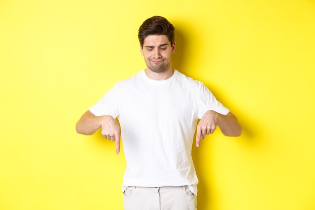 흰색 티셔츠에 회의적인 젊은 남자, 가리키는 화가 아래를 내려다 보면서, 승인하지 않고 제품을 싫어하고, 노란색 배경 위에 서 있습니다.