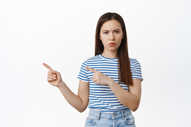 Skeptical young woman frowning, squinting in disbelief, pointing fingers left at sale logo, showing smth suspicious, dislike banner, complaining, standing over white wall