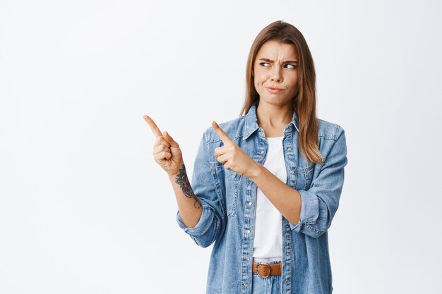 Skeptical young woman having doubts, smirk and frowning dissatisfied at logo, pointing and looking at upper left corner, standing against white wall